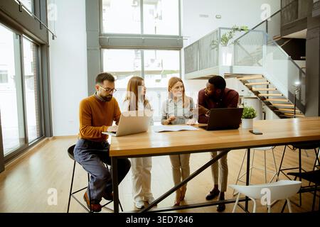 Four people sit and stand around a wooden table in a large office area. They share ideas while looking at laptops and documents. Windows let in sunlig Stock Photo