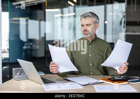 Senior businessman with gray hair and beard holding documents and reviewing paperwork at his desk, working on a laptop in a contemporary glass office setting Stock Photo