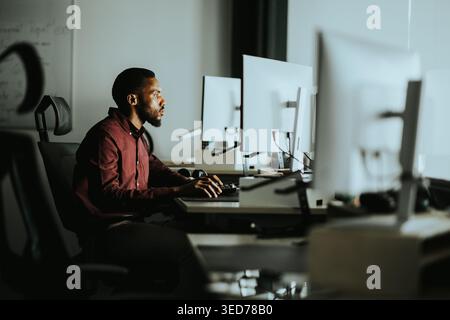 Late in the evening, a focused man sits at a sleek desk, concentrating on his tasks amidst bright computer screens. Stock Photo