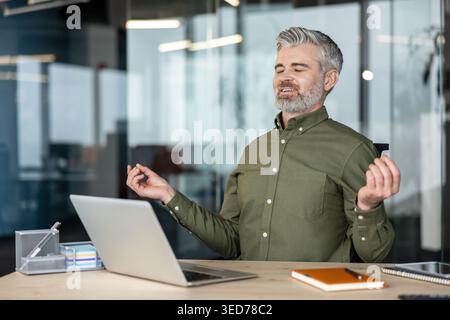 Mature businessman closing eyes and meditating at his desk in a blur-background office, practicing mindfulness and stress relief during a work break by finding zen moment Stock Photo