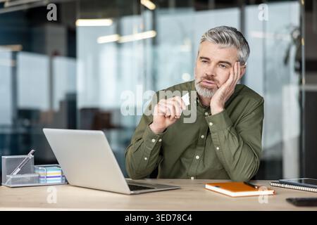 Mature man with gray hair looking away from his laptop, holding a crumpled piece of paper, reflecting on a problem or feeling bored at his modern office desk Stock Photo