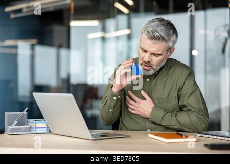 Mature man suffering from an asthma attack and breathing difficulties while sitting at his office desk, using an inhaler for relief and urgently needing treatment Stock Photo