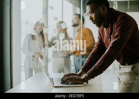 A man is using a laptop at a table in a bright office. In the background, a group of people talks and works together on a project. They are focused on Stock Photo