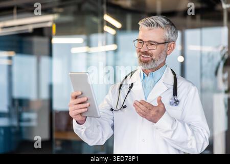 Doctor wearing a lab coat and stethoscope, smiling while having a telehealth video call, communicating with a patient using a tablet, offering remote medical assistance and advice Stock Photo