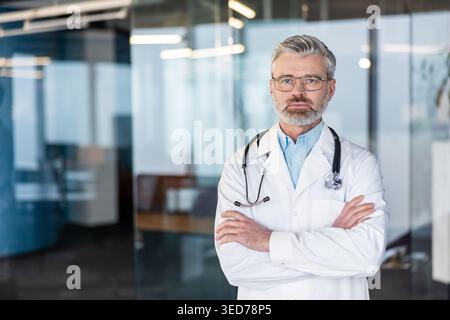 Experienced senior male doctor standing with arms crossed in white lab coat and stethoscope, confident, trustworthy medical professional in clinic or hospital setting, looking at camera Stock Photo