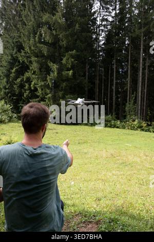 Young man catching small white drone by hand on green mountain meadow near spruce forest. Modern technology usage in nature, summer outdoor activity, Stock Photo