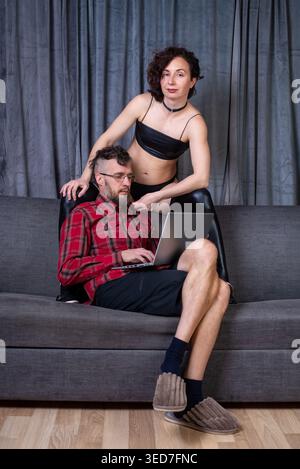 Man working on a laptop on a sofa while his partner in a black crop top watches playfully, creating an intimate home office moment Stock Photo
