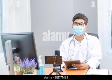 Portrait of a doctor sitting at a desk in an office with assorted digital devices wearing a face mask and laboratory coat Stock Photo