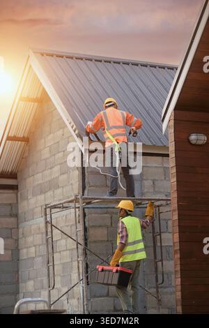 Side view of a Construction worker installing metal sheets on a roof and a coworker standing on scaffolding below, Thailand Stock Photo