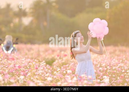 Portrait of a smiling young woman in a summer dress standing in a field of pink flowers holding an oversized floral pinwheel Stock Photo