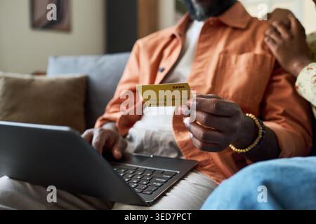 Black man holding bank card and using laptop while preparing trip booking at home, representing online travel payment and budgeting. Ideal for tourism, fintech, ecommerce campaigns Stock Photo