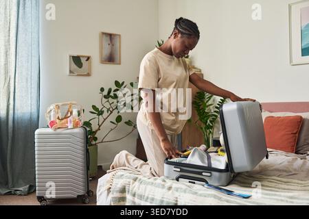 Black young adult woman packing suitcase on bed, preparing for trip and organizing travel essentials at home. Useful for vacation planning, tourism, travel service marketing Stock Photo
