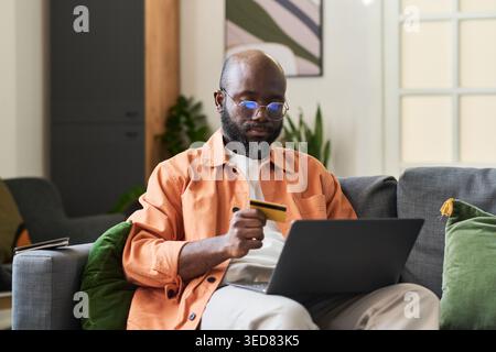 Medium shot of Black middle aged man holding credit card and using laptop while booking trip from home, planning travel purchase. Useful for tourism marketing, online payment Stock Photo