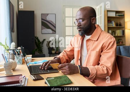 Black middle aged man booking travel on laptop while holding credit card and passport, preparing for trip. Ideal for tourism marketing, online booking, travel finance Stock Photo