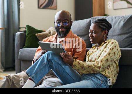 Black couple sitting on floor and using tablet while planning trip at home, discussing bookings and travel budget. Ideal for vacation planning, online booking, tourism marketing Stock Photo