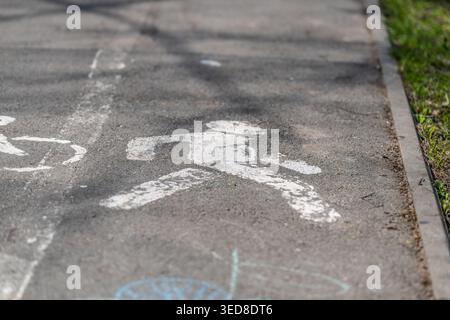 White pedestrian figure on shaded walkway, chalk doodles near edge and soft tree shadows crossing path, curb boundary and grass verge visible, invitin Stock Photo