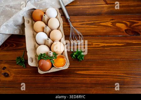 Raw eggs in carton with parsley and whisk on rustic wood. Stock Photo