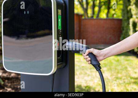 Hand Holding EV Charging Plug Outdoors. Close-up of a hand holding an electric vehicle charging connector near a modern outdoor charging station in a Stock Photo