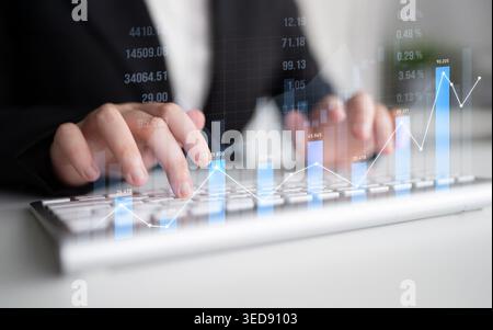 A business professional uses a keyboard while analyzing financial data trends. Overlaid graphs represent growth and market insights in a modern office Stock Photo