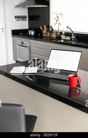 A laptop placed on a kitchen counter. There is a notebook and a pen nearby along with a red cup. The kitchen is modern and well organized. Stock Photo