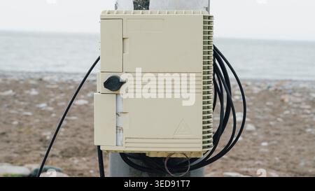 Beige fiber optic distribution box with black cables mounted outdoors near a beach Stock Photo