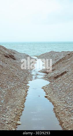 A small stream of water flowing through large piles of pebbles toward the sea Stock Photo