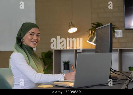 Woman wearing green gradient hijab typing on laptop with monitor and notebook at office desk Stock Photo