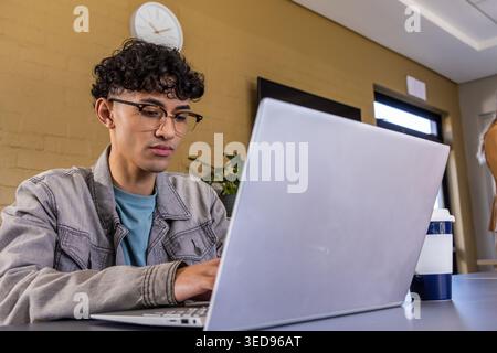 Man wearing denim jacket, glasses working on laptop at table in shared workspace with travel mug Stock Photo
