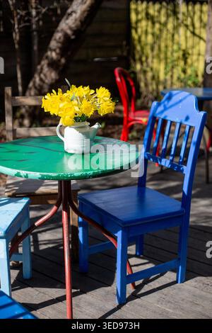 Yellow daffodils in a vase on a green table surrounded by colorful chairs in a sunny outdoor setting Stock Photo