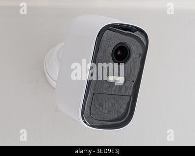 A close-up shot of a white smart home security camera installed on a white wall. The camera features a sleek design with a black glass face, integrate Stock Photo