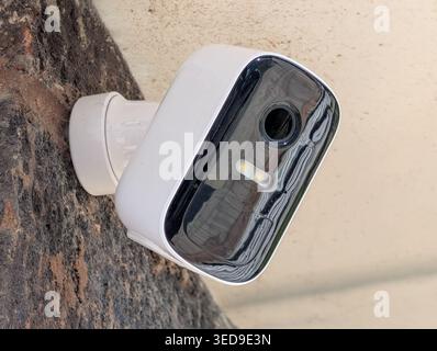A close-up shot of a white smart home security camera installed on an exterior brick wall. The camera features a sleek design with a black glass face, Stock Photo