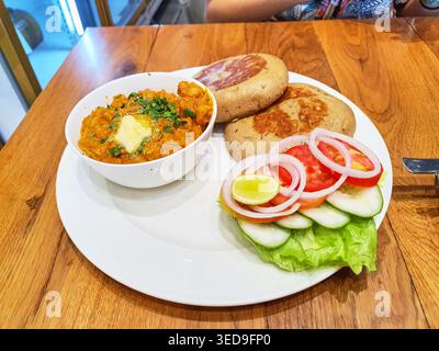 A white ceramic plate holds a traditional Indian meal served on a wooden restaurant table. The dish includes a white bowl filled with orange-colored Stock Photo