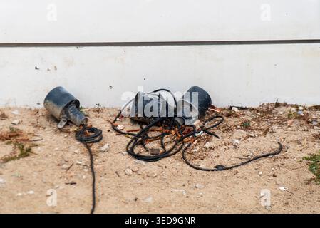 The image shows a tangled cluster of electrical cords and wires discarded on a dirt ground near a white wall, creating a cluttered outdoor scene. Stock Photo