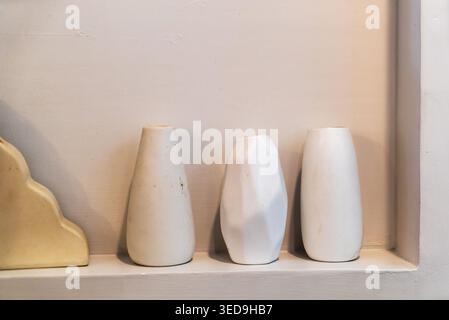 A minimalist collection of three white ceramic vases with different shapes, displayed on a shelf against a neutral background, showcasing simple elega Stock Photo