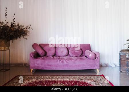 Purple sofa sits softly against white curtains. Rug with intricate patterns lies beneath. Lush plant adds earthy charm to the corner. Minimalist style Stock Photo