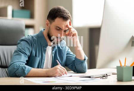 Focused young businessman sitting at desk in office and writing in notepad Stock Photo