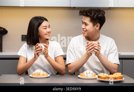 Asian Couple Having Breakfast Sitting In Kitchen On Weekend Morning Stock Photo