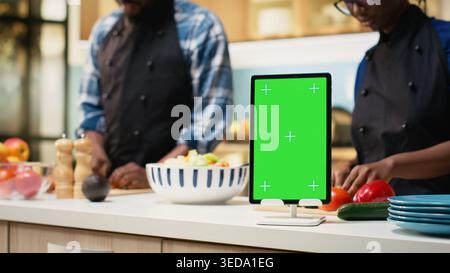 Vertical chroma key tablet with lovers doing meal prep for a salad, preparing food at the kitchen counter. Couple enjoying time together in the kitchen, cooking with aprons and having fun. Stock Photo