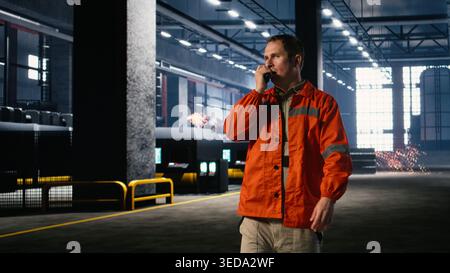 Heavy industry employee speaks into walkie talkie device in warehouse, ensuring safe workflow and manpower coordination. Communication during construction and fabrication operations. Stock Photo