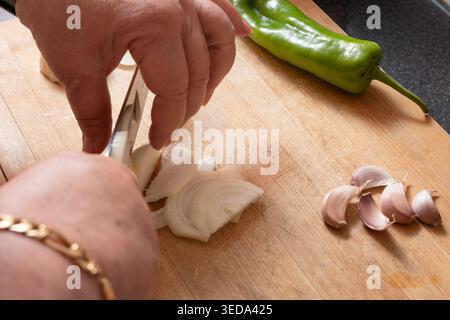 Hands slicing fresh onion on a worn wooden cutting board with garlic and a green pepper Stock Photo