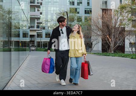 A cheerful couple strolls hand in hand, delightfully holding colorful shopping bags as they share smiles and laughter, making the most of their Stock Photo