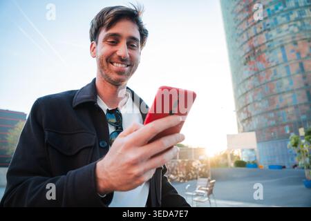 Joyful man smiling as he checks notifications on his smartphone, displaying excitement and engagement with technology in a lively outdoor atmosphere Stock Photo