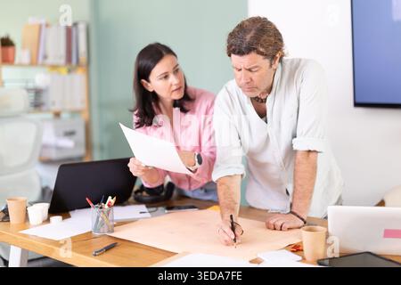 Middle-aged colleagues discussing construction plan leaning over office table Stock Photo
