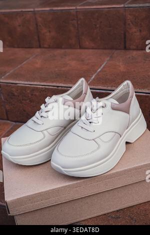 Minimalist eco-friendly white sneakers placed on recycled cardboard box, clean design for sustainable fashion concepts. New unbranded beige sneakers footwear mockups and modern product displays. For branding, e-commerce and packaging design Stock Photo