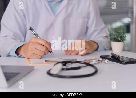 Close up doctor hand writing prescription on clipboard with stethoscope on desk. Professional physician filling medical report and insurance form in h Stock Photo