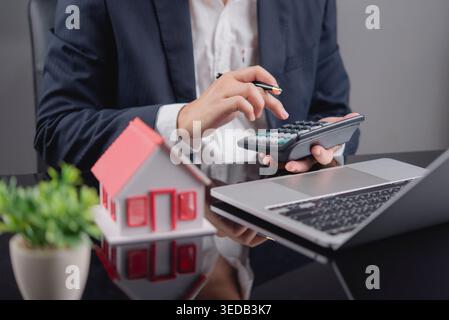 Businessman using calculator for calculating finance on table with house model. Real estate, tax, and property investment concept. Stock Photo
