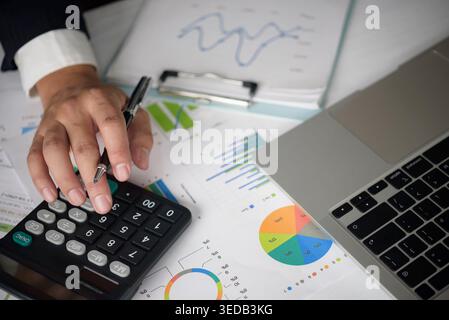 Close up businessman hand using calculator and writing on financial report with charts and laptop on desk. Professional accountant analyzing budget an Stock Photo
