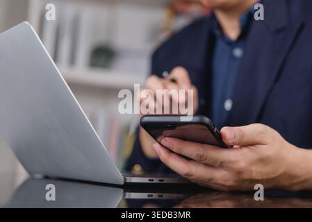 Close-up of a businessman's hands holding a smartphone and a pen while working on a laptop computer at an office desk. Concept of multitasking and mod Stock Photo