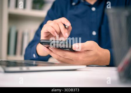 Close up businessman or freelancer hand holding a smartphone and a pen while sitting at a white desk with a laptop. checking email, mobile banking app Stock Photo