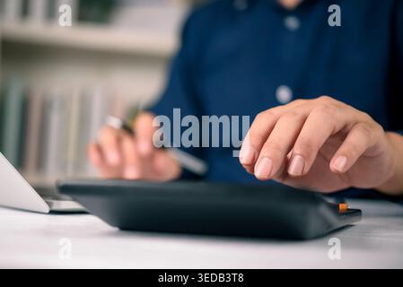 Close up of a man hand using a black calculator on a white wooden table with a blurred bookshelf background. Concept of financial planning, calculatin Stock Photo
